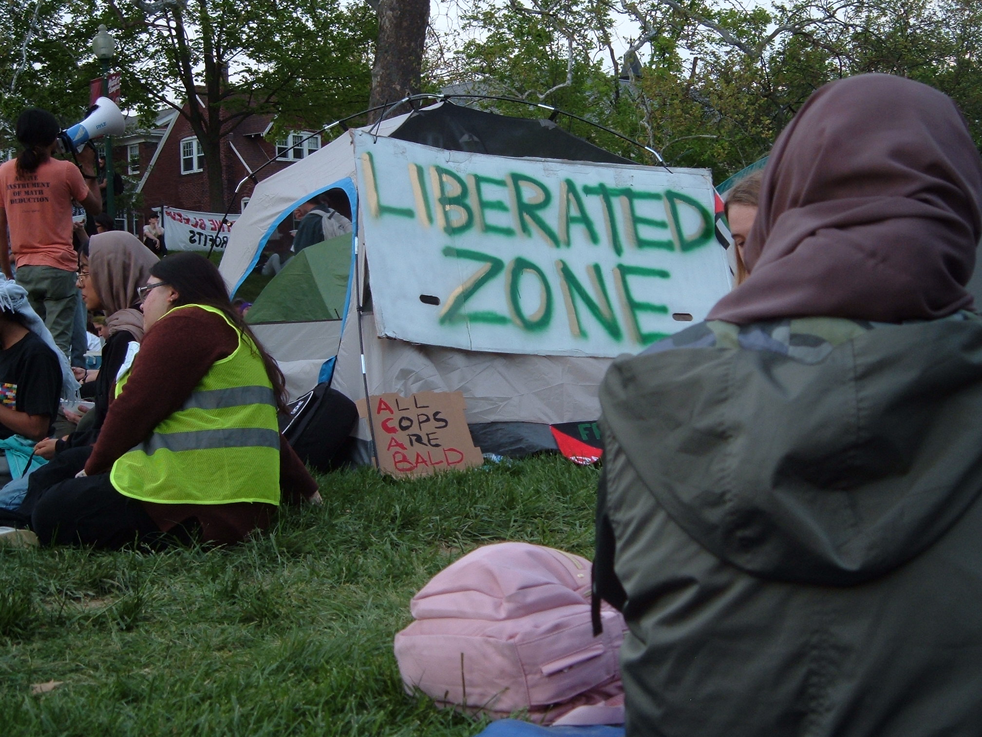 Protestors sit and chat on the grass in the encampment. A sign that says 'Liberated Zone' in green and gold letters is affixed to a tent. Below that is a sign that reads 'All Cops Are Bald' in red and black letters.