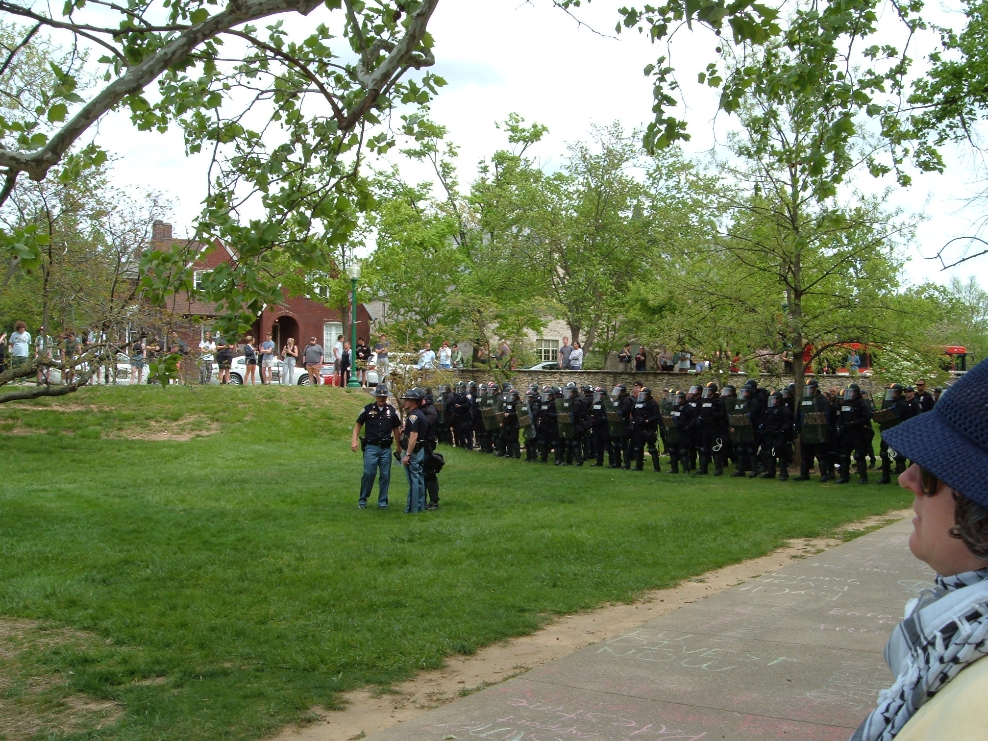 Indiana State Police in riot gear line up on the green grass of Dunn Meadow, waiting for the order to begin the raid. The commanding officers converse in a small group in front of the line.