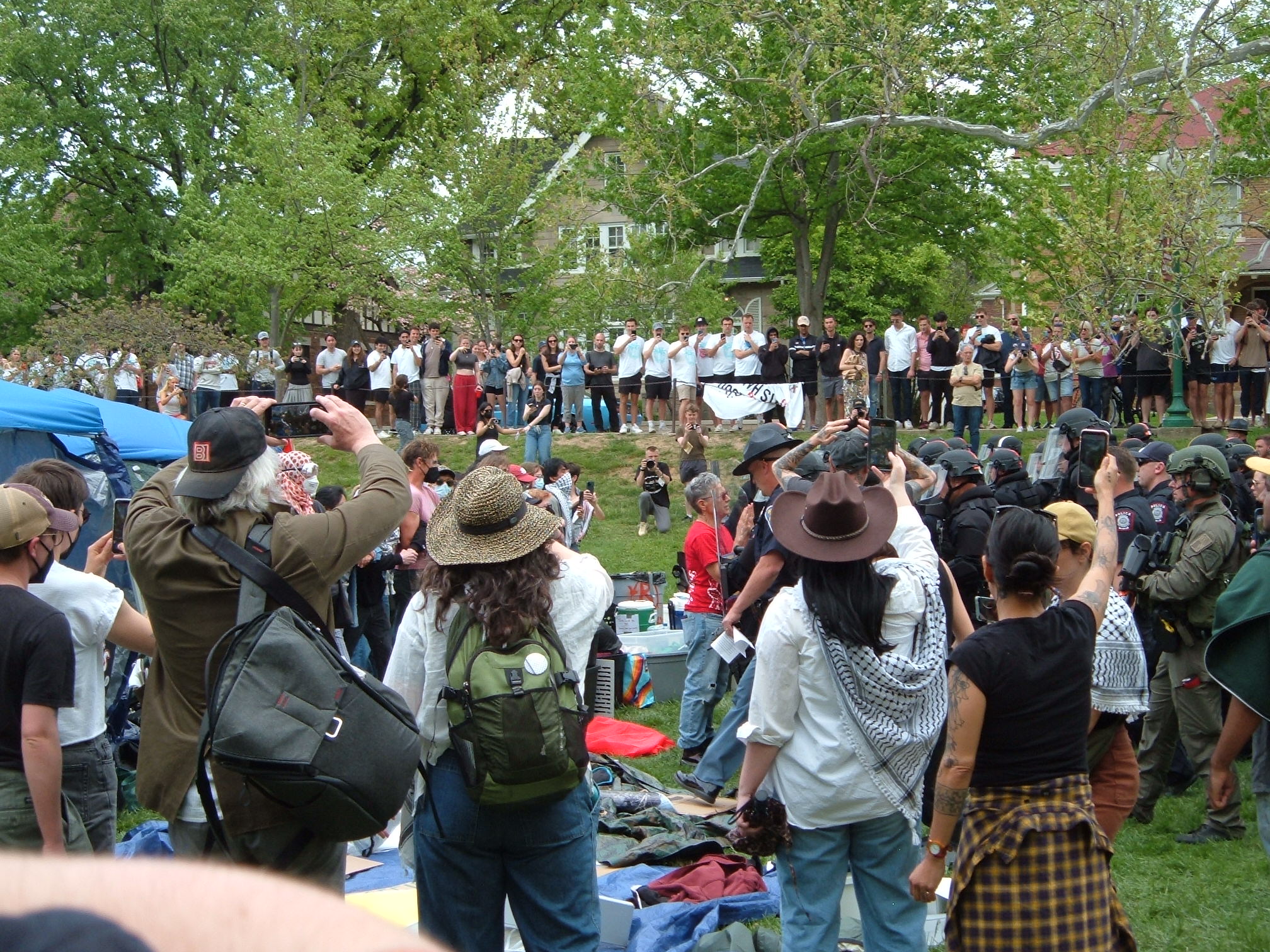 Indiana State Police in riot gear move towards the encampment. A woman in a red shirt begs the commanding officer to stop. People film the march of the riot cops. Above Dunn Meadow on the sidewalk, the crowd of observers has grown.