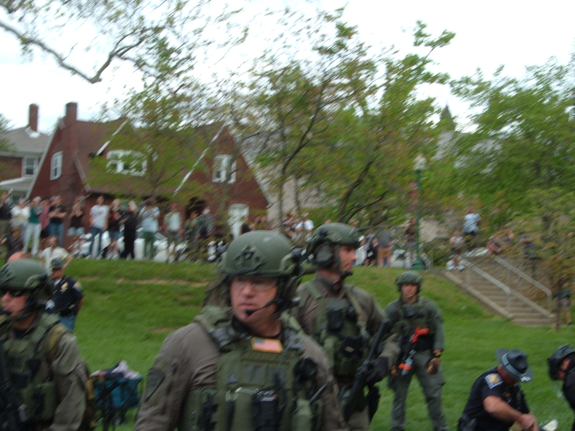 A blurry photo of the SWAT unit with their fingers on the triggers of their weapons stand around as the riot police conduct arrests. Behind them on the sidewalk is a group of onlookers, filming.