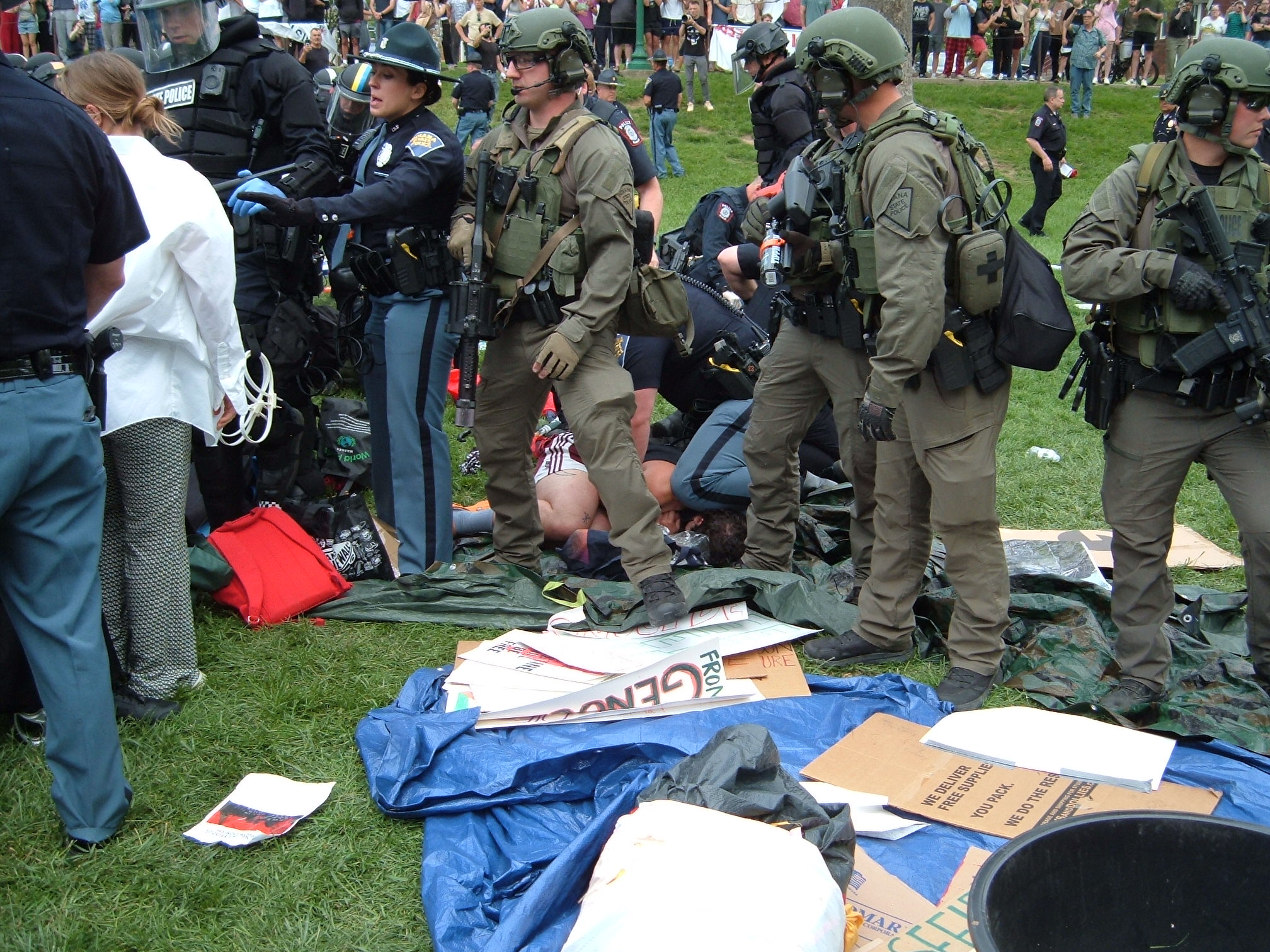 Indiana State Police in riot gear arrest a protestor. One officer kneels on their head and neck as police wrestle the protestor's hands into zip tie handcuffs. The destroyed tent and signs litter the area.