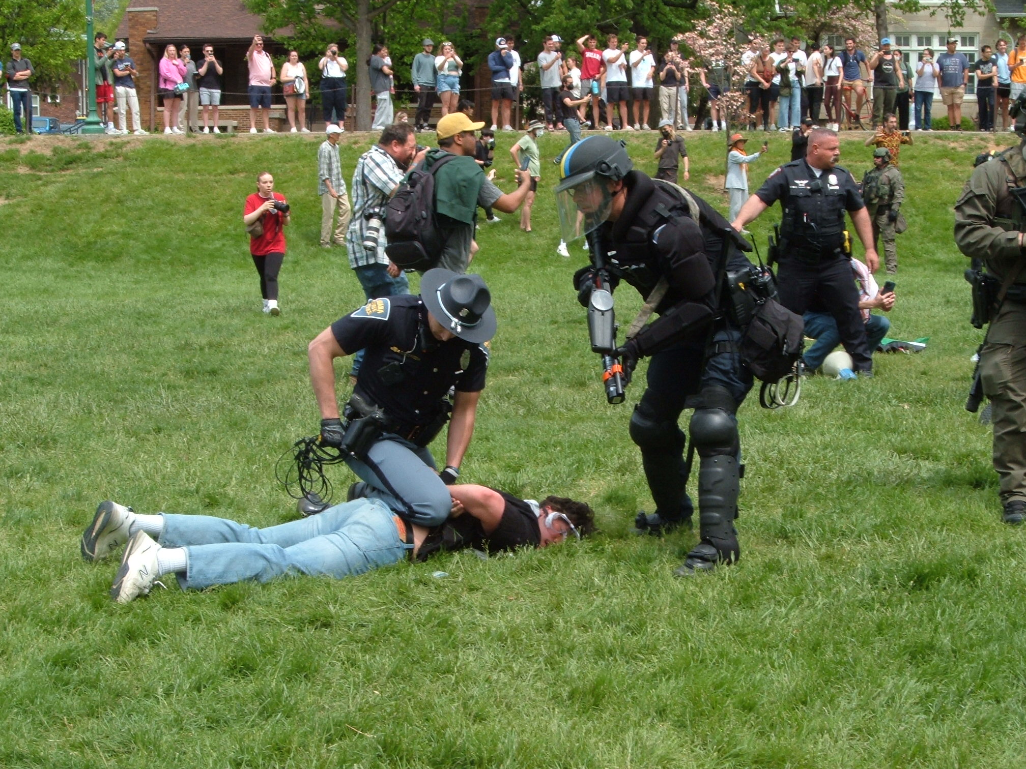A policeman in riot gear stands over a protestor lying facedown in the grass, handcuffed. The policeman aims his gun loaded with rubber bullets near the protestor. Another officer sits on them restraining them.