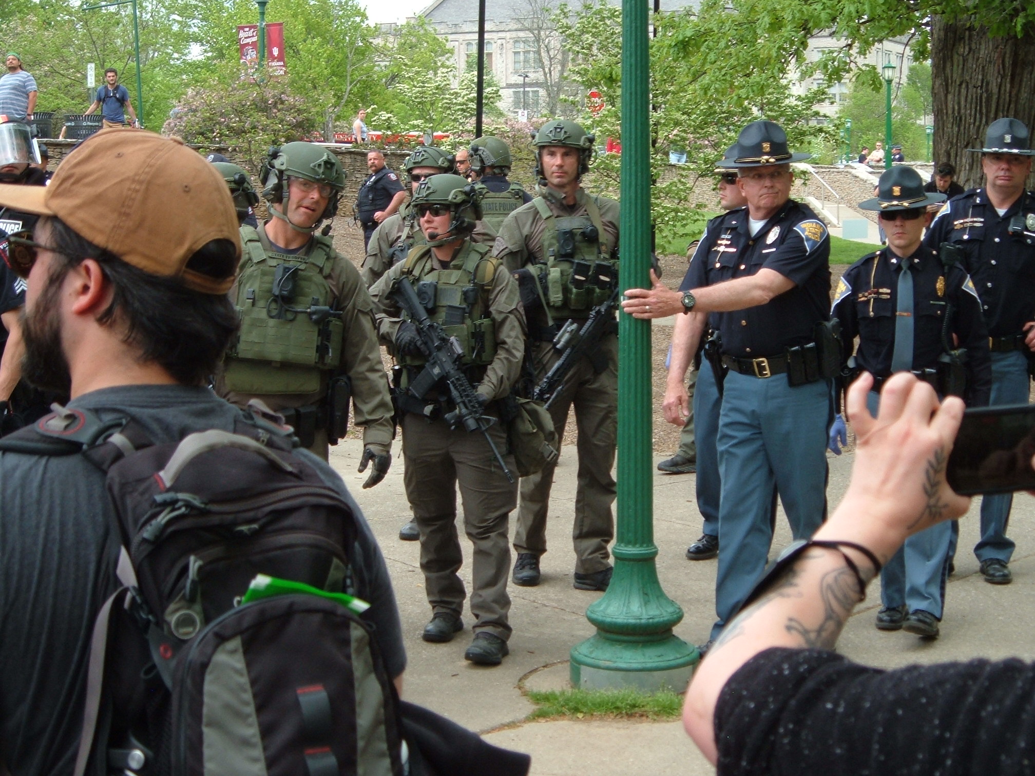 The commanding officer holds a green lamp post that several police nearly tripped over in the retreat. On one side of the frame, the SWAT unit with guns exchanges words. On the other side, Indiana State Police officers in uniform observe the crowd.