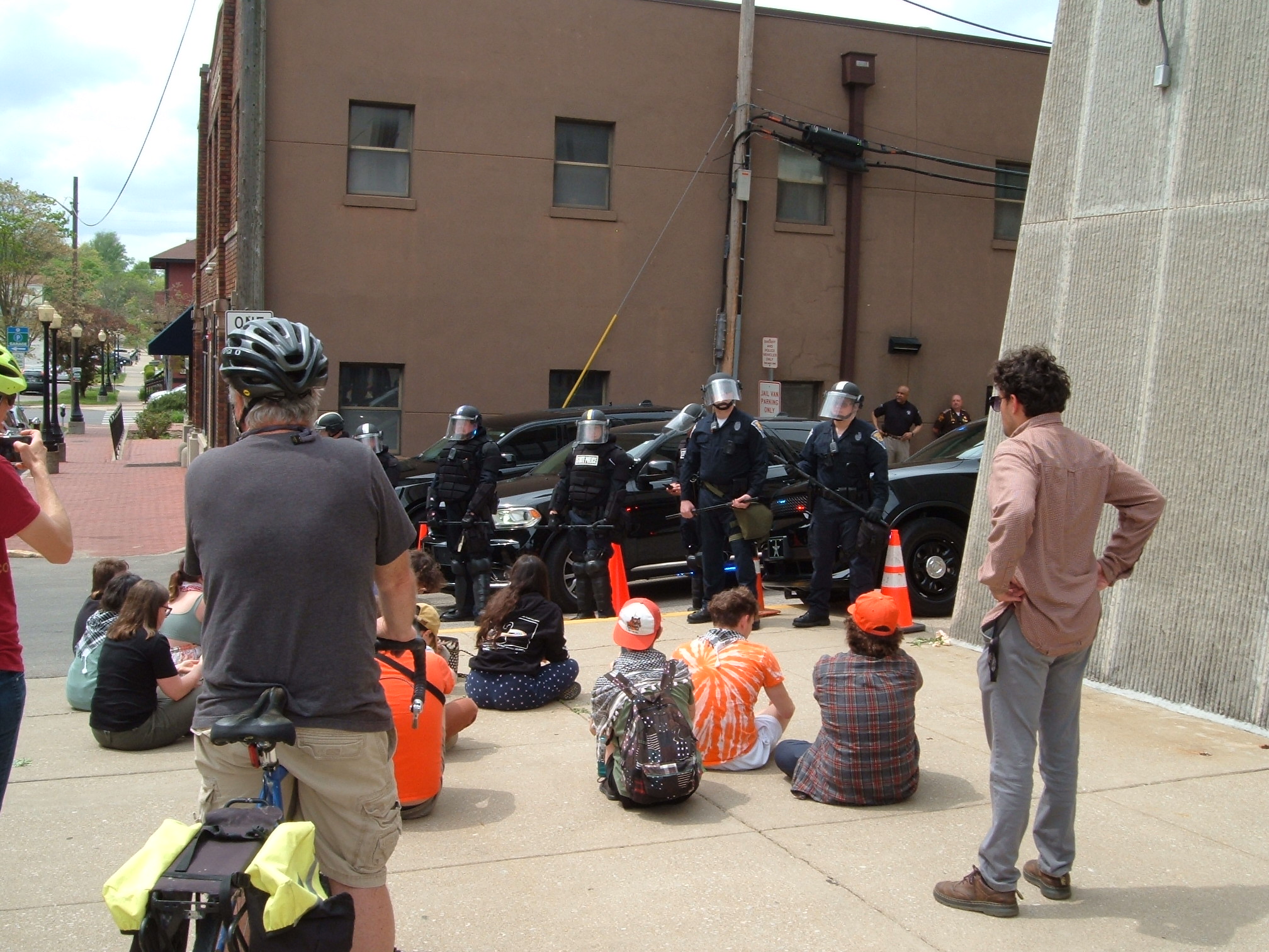 Several protestors sit on the sidewalk in front of a unit of riot police guarding the County Jail.